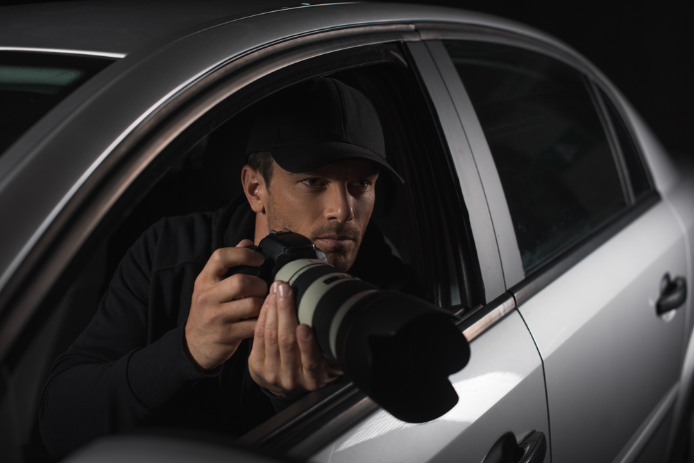 Man with a baseball cap on sat in a car with a long lens camerca